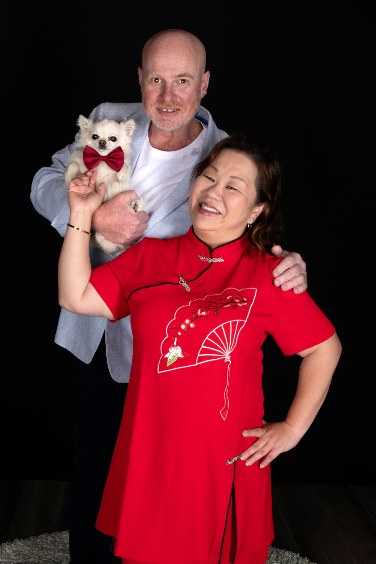 Couple laughing together, the husband holding their small white chihuahua in a red bow tie beside his wife in a red mandarin collar dress
