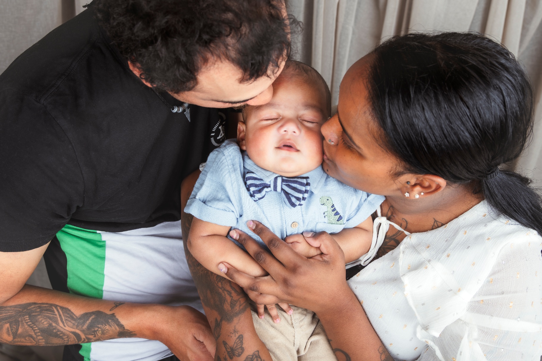 Mum and dad each kissing their sleeping newborn son, dressed in a blue shirt and bow tie, held between them