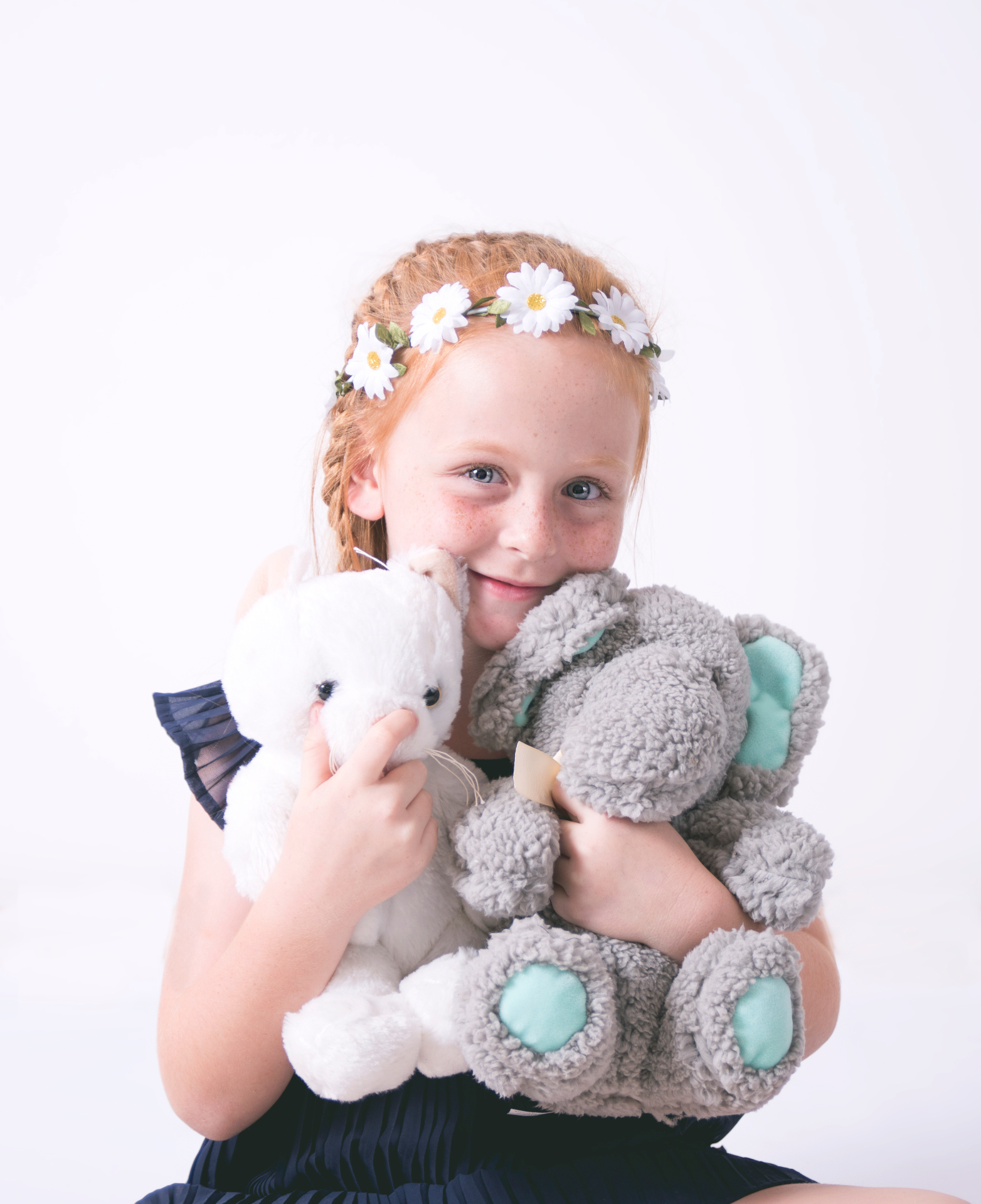 Young red-haired girl with a daisy flower crown cuddling a white rabbit toy and grey elephant plush in a bright studio portrait
