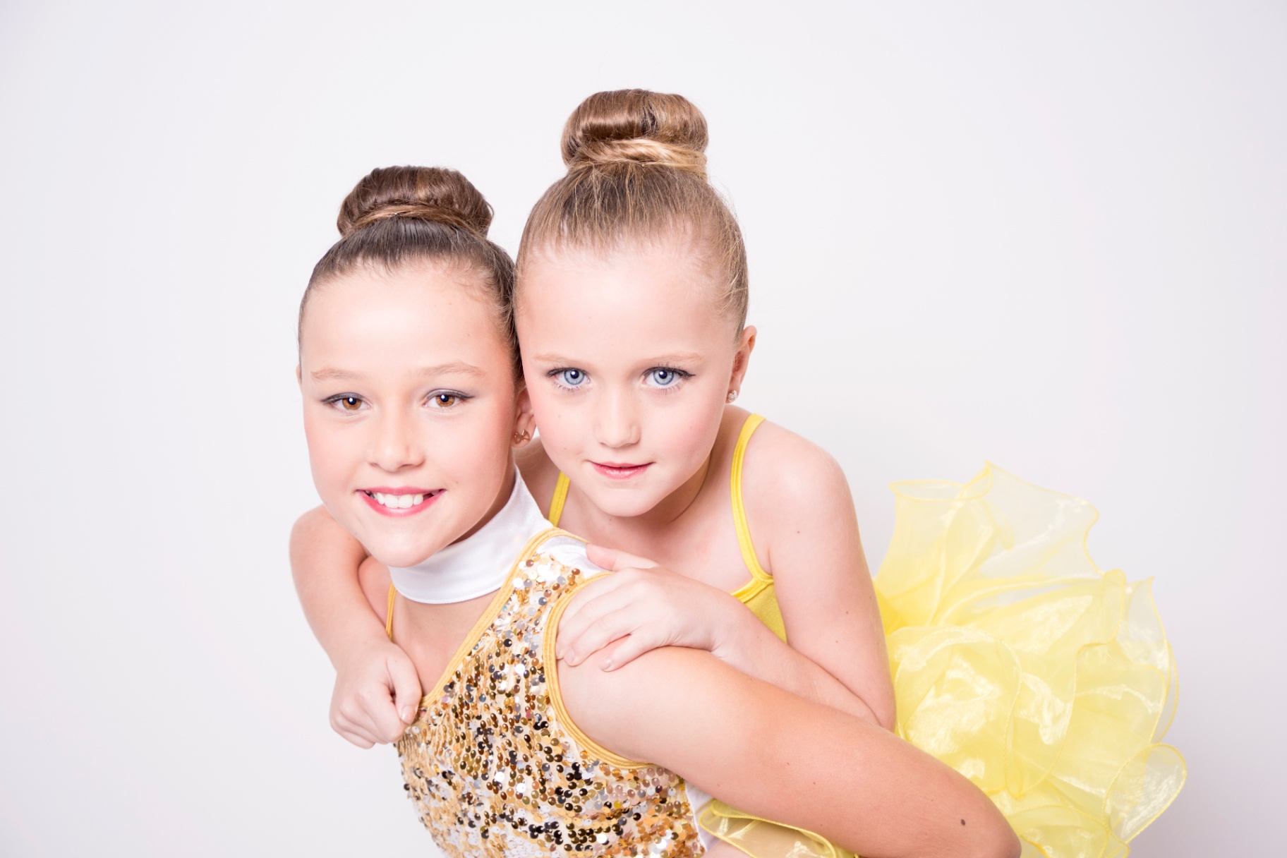 Two young sisters in gold and yellow dance costumes with sleek buns, cheek to cheek in the studio