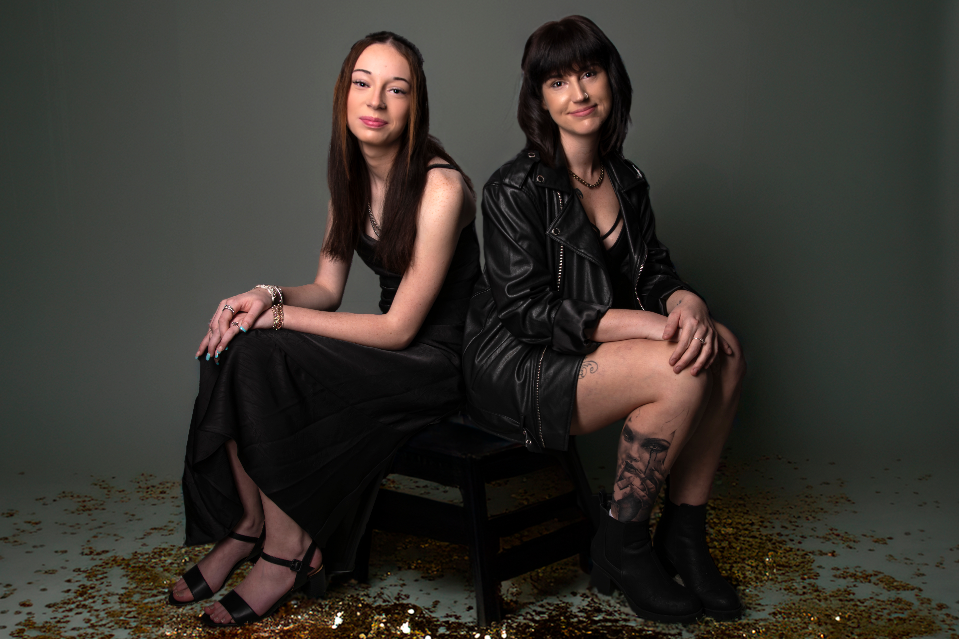 Two young women in black outfits posed back to back, seated in a modern concept portrait with scattered gold confetti