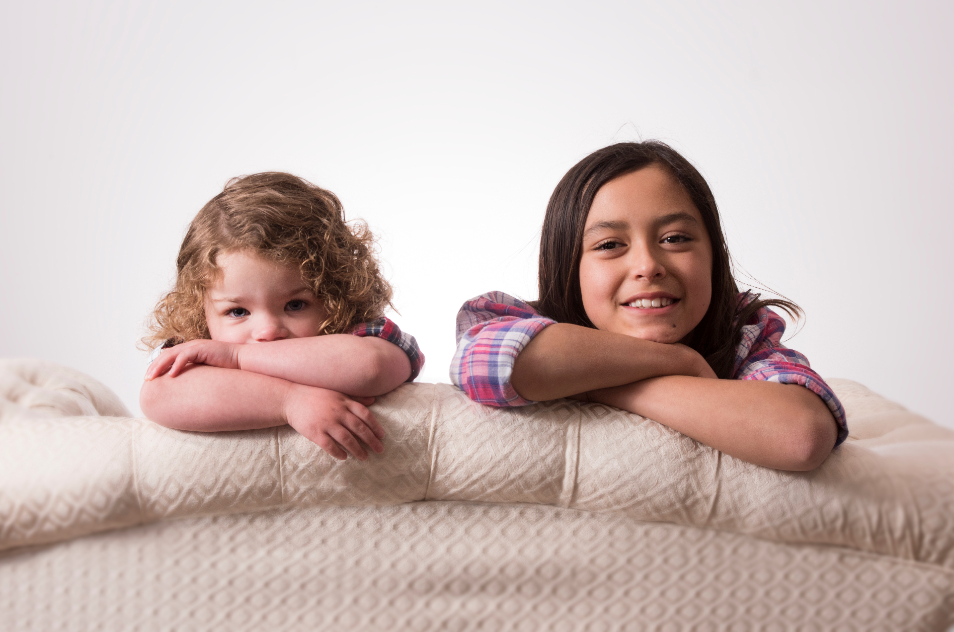Two young sisters resting their arms on a cream tufted mattress, smiling in matching pink check shirts