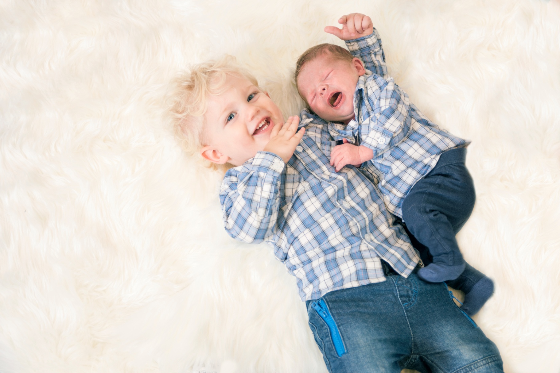 Big brother grinning beside his yawning newborn baby brother, both in matching blue check shirts on a white sheepskin