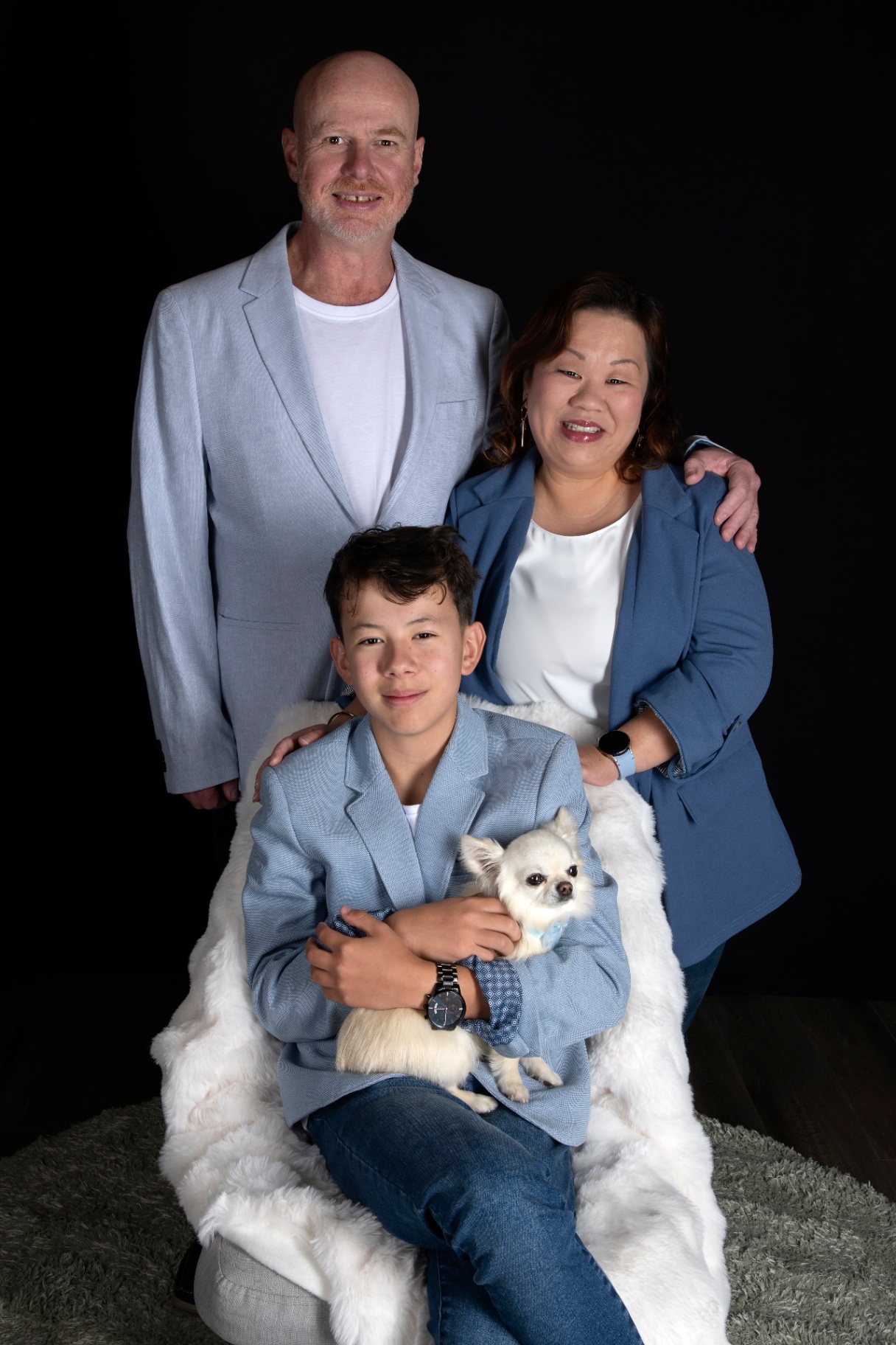 Family of three seated together with their small white chihuahua, dressed in coordinated blue tones on a soft sheepskin