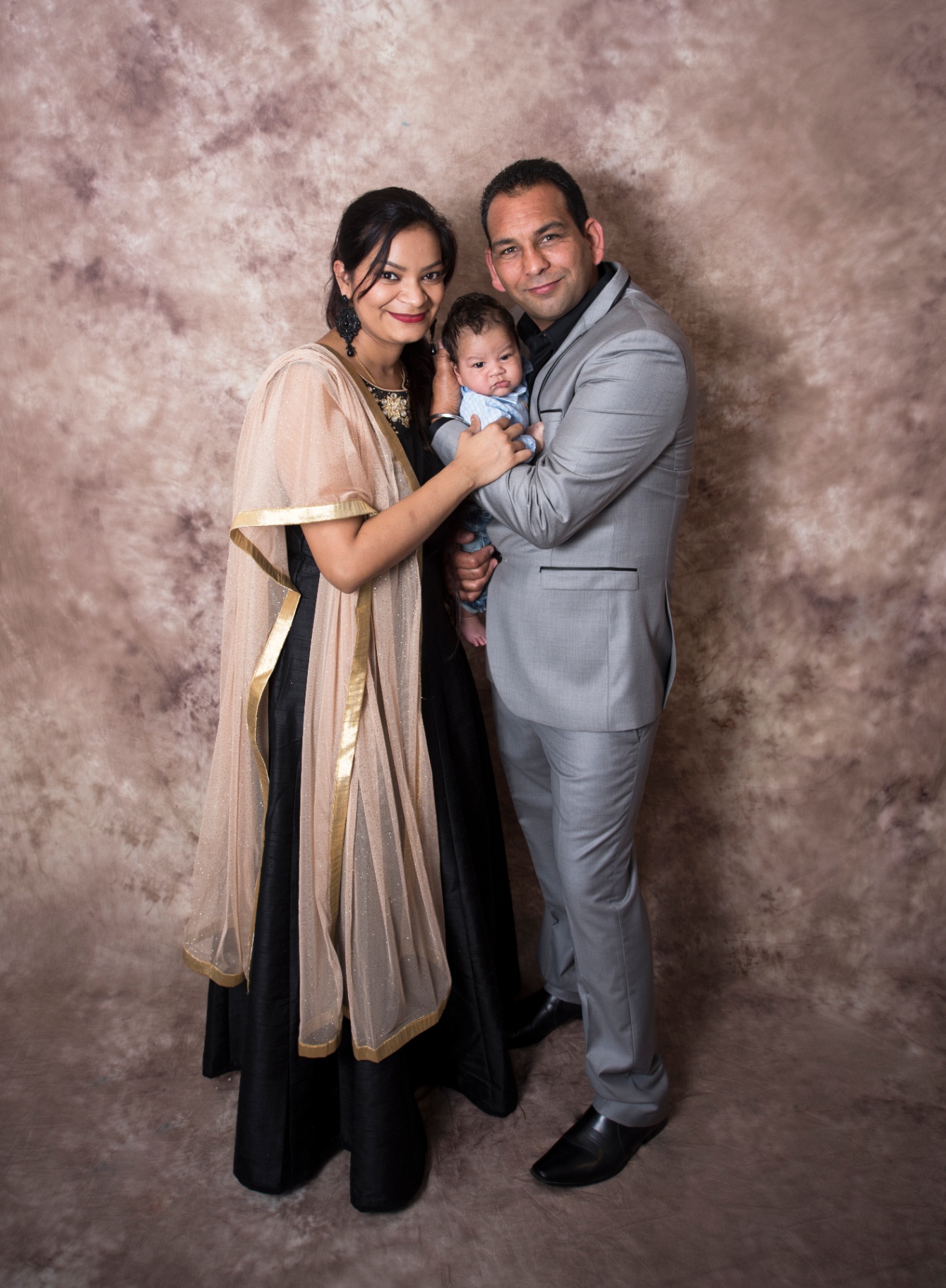 Mum and dad in elegant formal wear cradling their newborn son together against a warm mauve backdrop