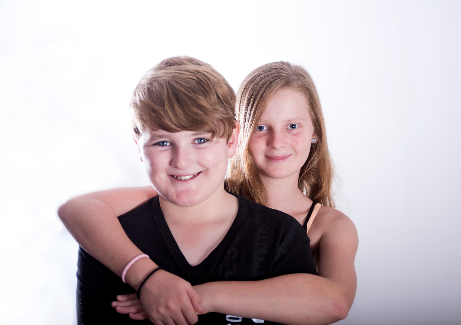 Brother and sister in black tops, the older sister hugging her younger brother from behind, both smiling softly