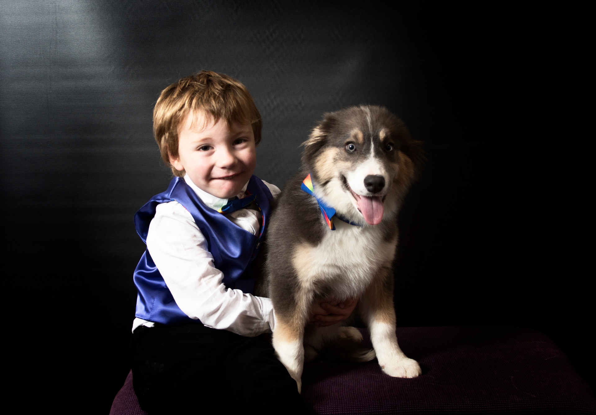 Young boy in a blue satin vest sitting beside a fluffy dog with bright blue eyes, both wearing rainbow bow ties