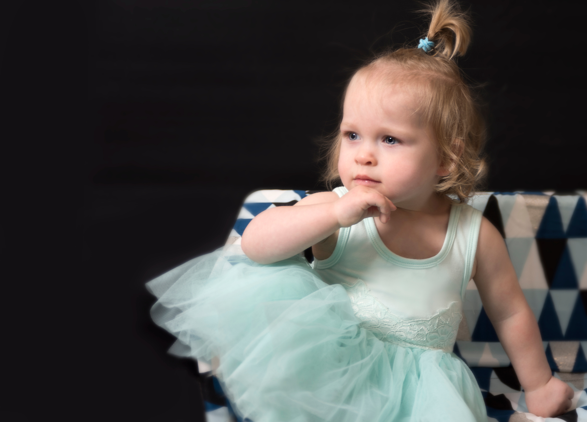 Thoughtful toddler girl in a pale aqua tulle dress, seated on a geometric armchair against a black backdrop
