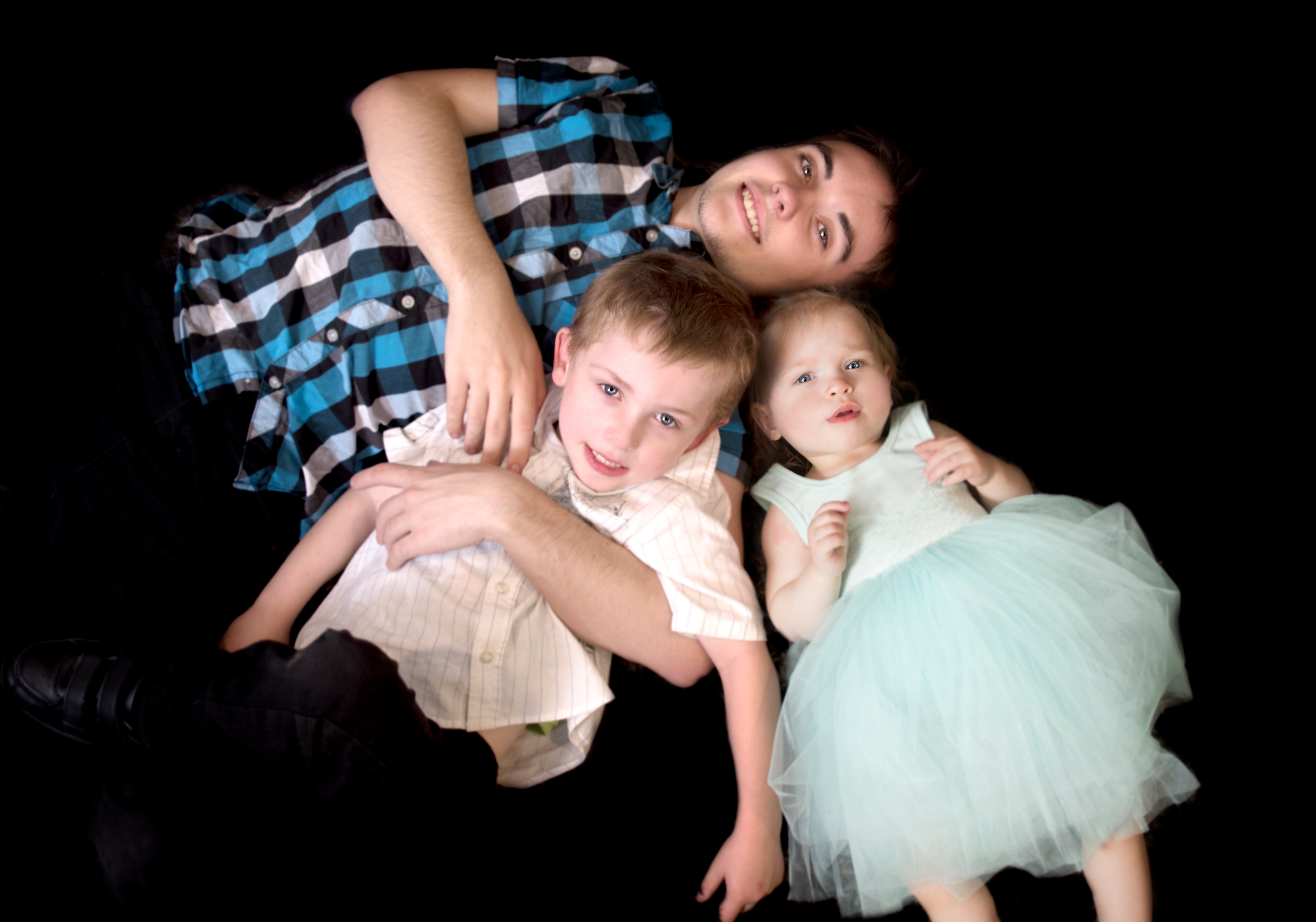 Dad lying on a black backdrop with his young son and toddler daughter tucked in close, all smiling up at the camera