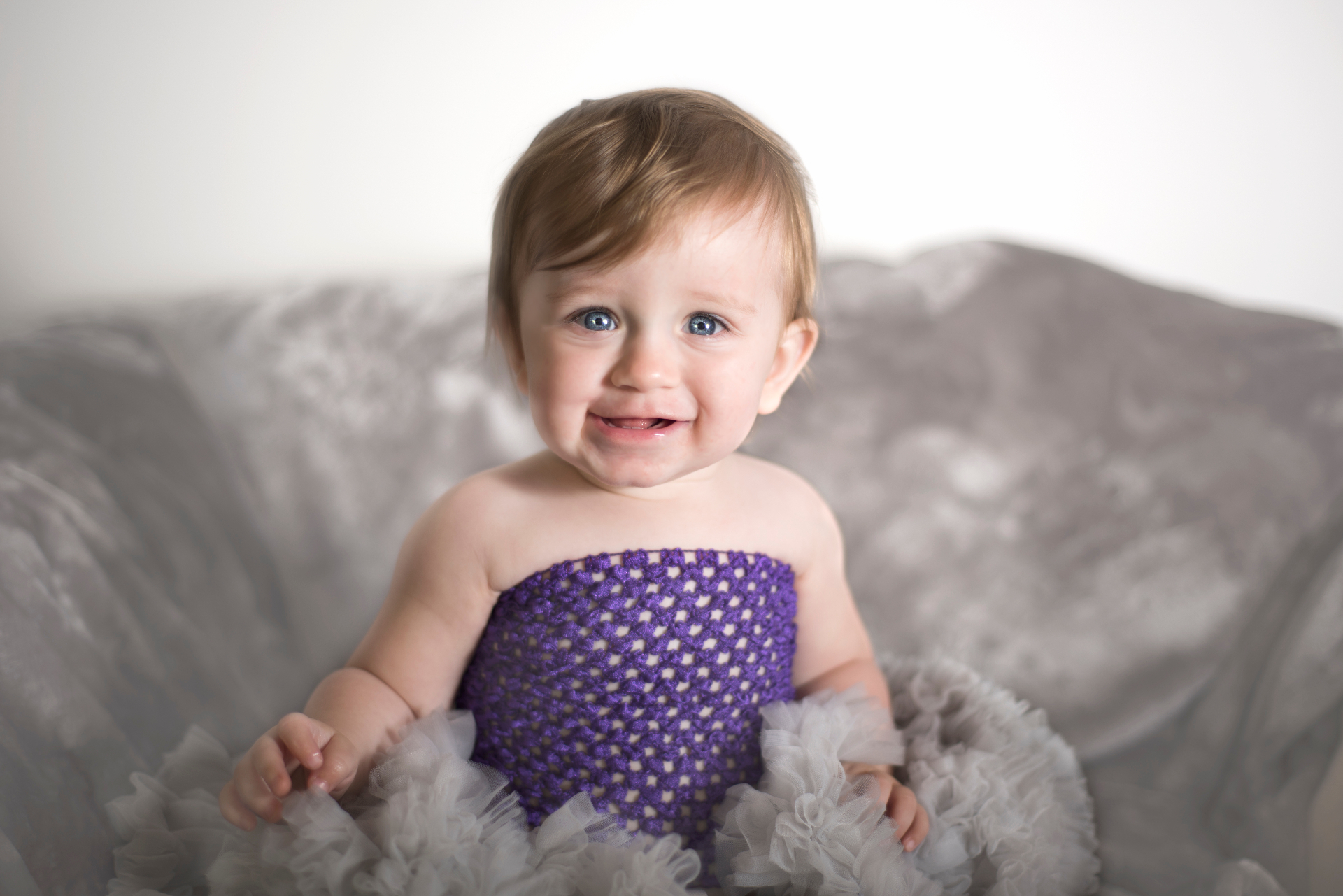 Smiling blue-eyed baby girl Jakyla in a purple tulle dress on a soft grey velvet couch