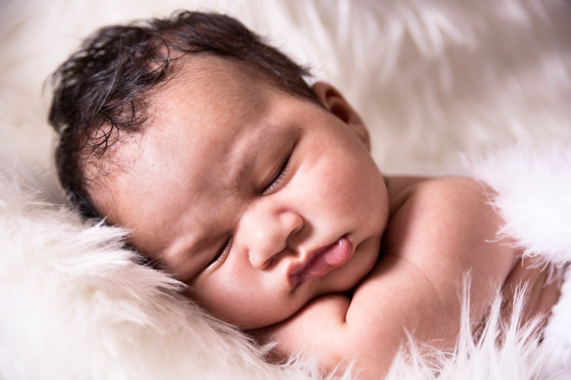 Sleeping newborn Amandeep with dark hair, nestled into a soft white fur blanket