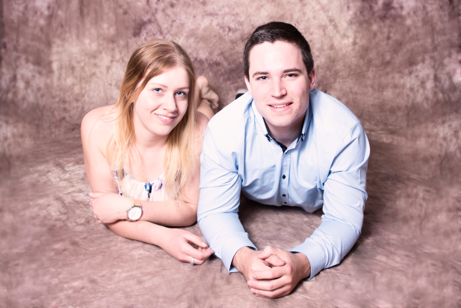 Young couple lying together on a soft mauve backdrop, resting on their elbows and smiling gently