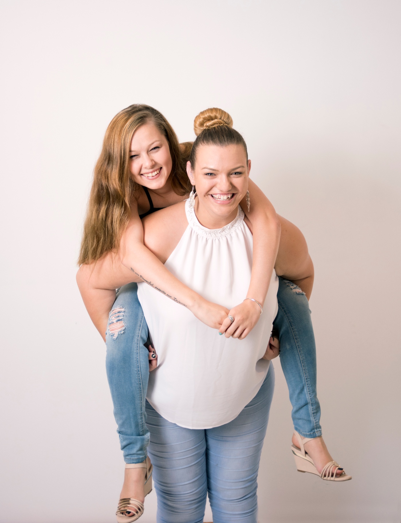 Two young women laughing together, one giving the other a piggyback in a bright studio portrait