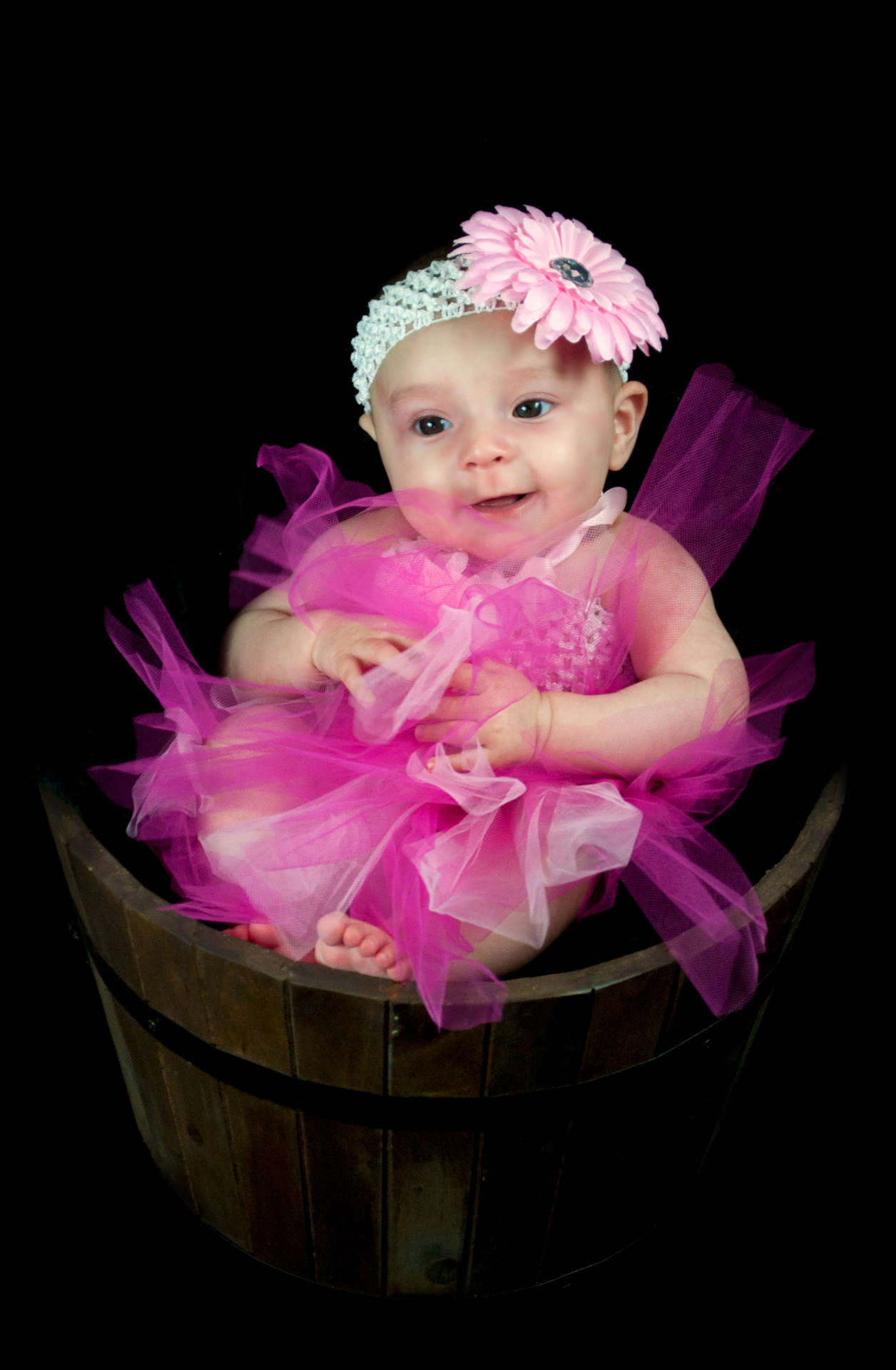 Smiling baby in a bright pink tulle tutu with a pink daisy headband, seated in a rustic wooden bucket against a black backdrop